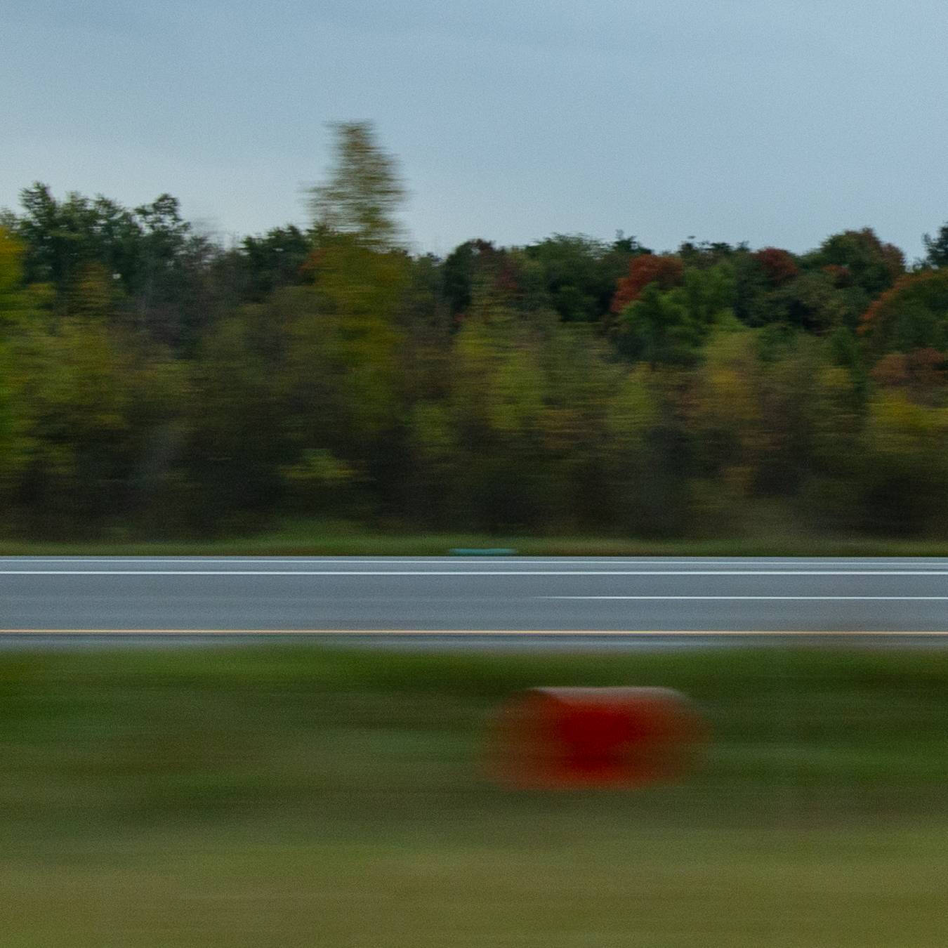 Blurry color photograph of the side of a road with trees and grass and a red mailbox.